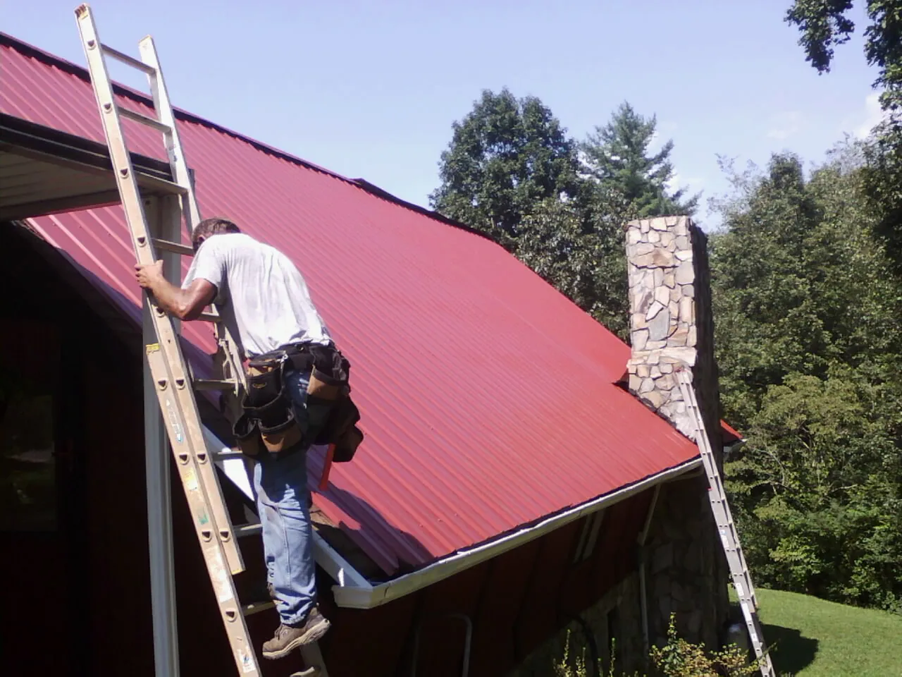 Roofer on a ladder beside a red metal roof and stone chimney — Soffit & Fascia Repair by Roofing Brothers in Cordova